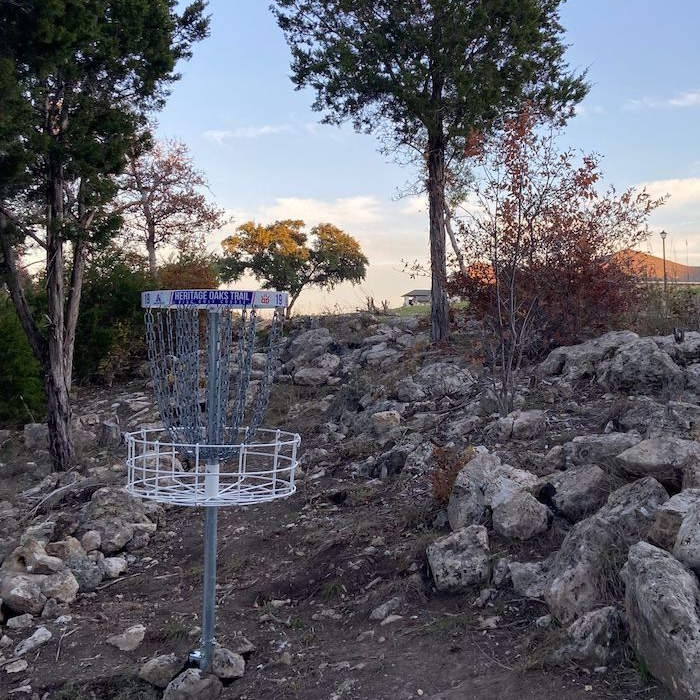 Heritage Oaks Disc Golf Course. There is a basket sitting on a path surrounded by rocks